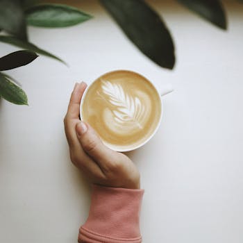 A cozy scene featuring a hand holding a latte with beautiful art, surrounded by green leaves on a white background.