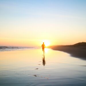 A lone silhouette of a person running along a serene beach at sunset, reflecting in the wet sand.
